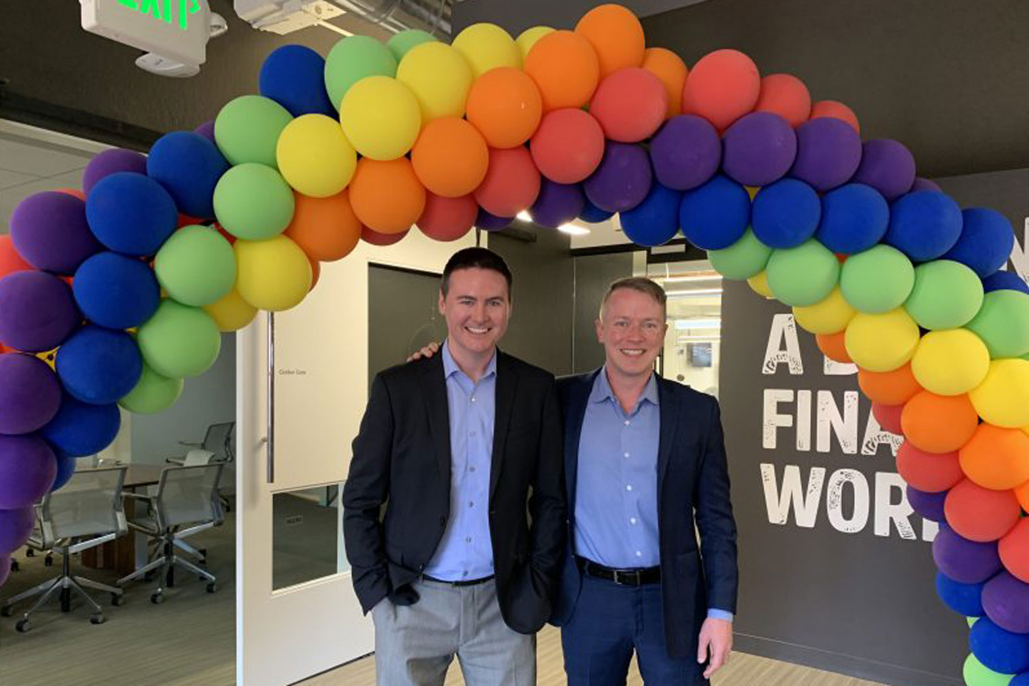 two men standing under rainbow balloon arch